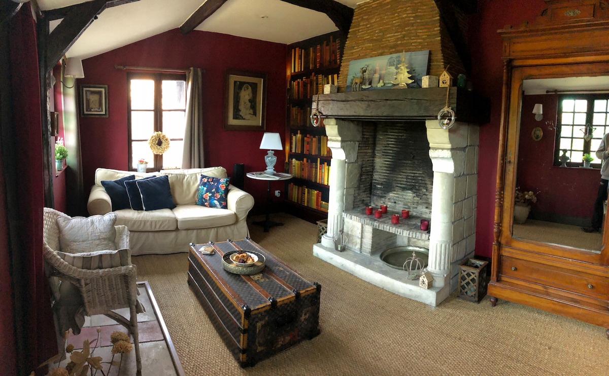 A cozy living area features a white sofa adorned with colorful cushions, complemented by a wicker chair. A rustic coffee table sits in front of a stone fireplace, with decorative items arranged on it. Shelves lined with books and artwork add character to the warm, inviting space.