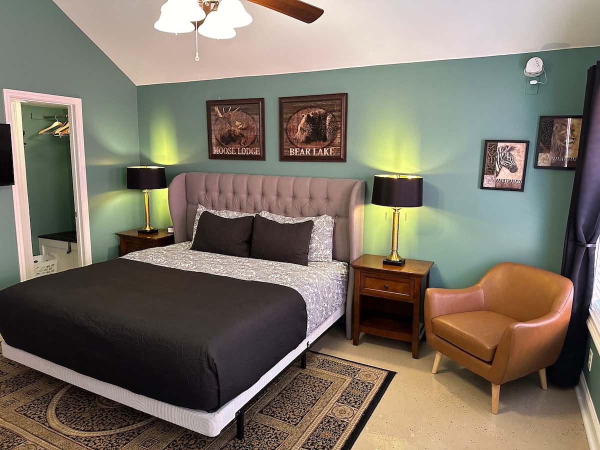 The bedroom features a spacious bed with a dark coverlet, complemented by decorative pillows. Two bedside tables with lamps are positioned beside the bed. A comfortable chair sits nearby, and nature-themed artwork adorns the walls. Soft lighting and a large window enhance the room's ambiance.