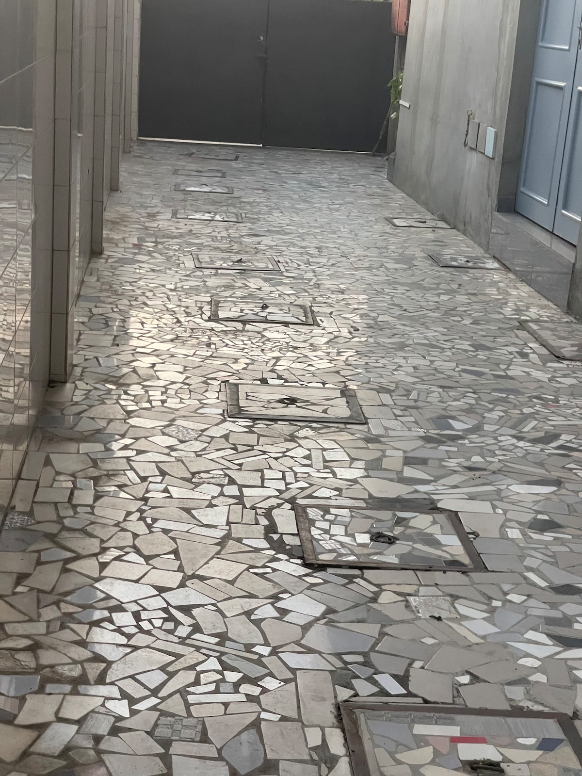A narrow pathway is lined with textured stone tiles, featuring a mosaic pattern. Manhole covers are evenly spaced along the ground. A solid gate is visible at the end of the walkway, which is framed by simple, concrete walls on either side.