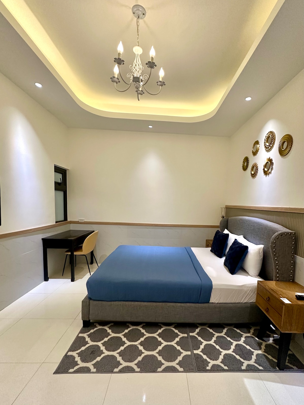 The bedroom features a cozy double bed with a blue blanket, complemented by two decorative pillows. A contemporary chandelier hangs from the ceiling, illuminating the space. A small desk with a chair is positioned against the wall, alongside decorative plates arranged above the bed.