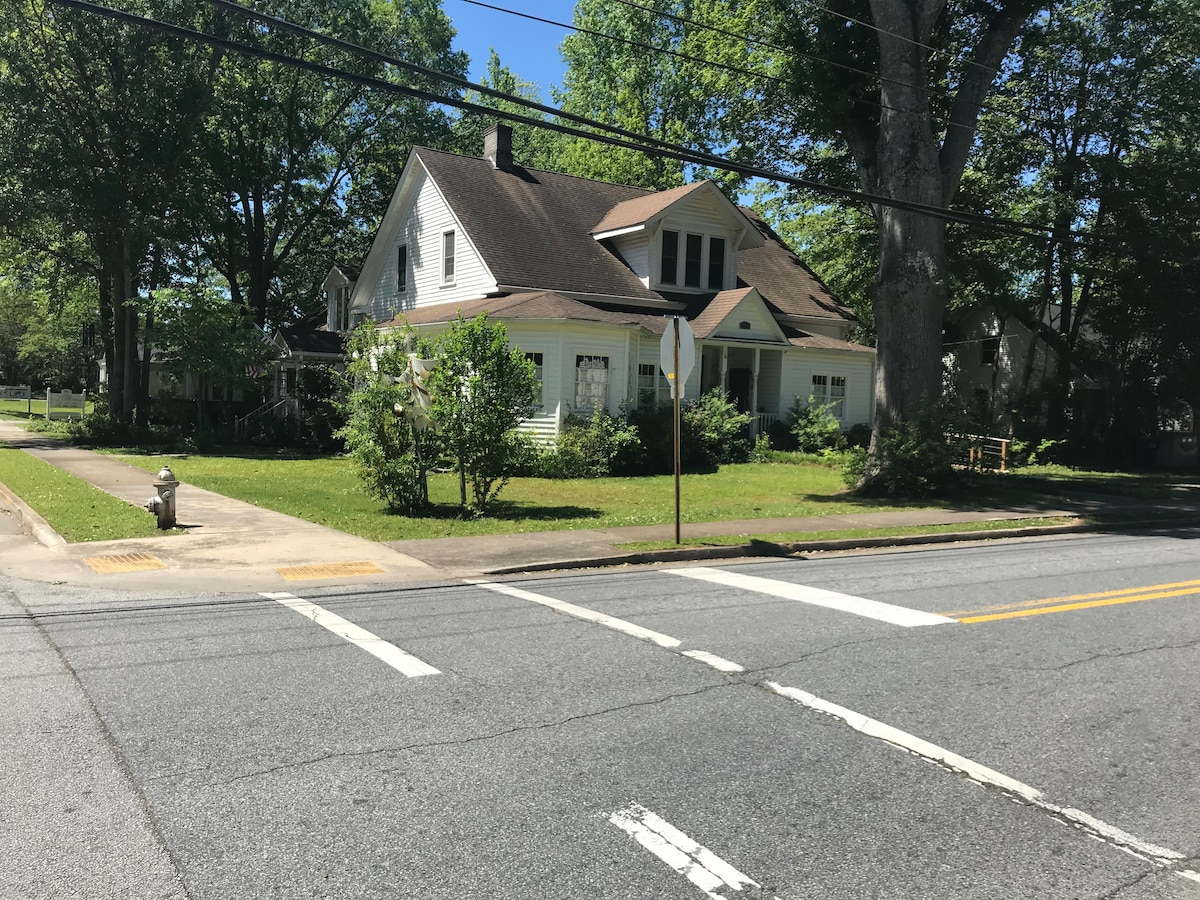 A spacious house with a sloped roof and large windows is surrounded by green lawns and trees. The structure is positioned at the corner of a street, with a clear view of the front yard and nearby sidewalk. A fire hydrant is visible in the foreground.