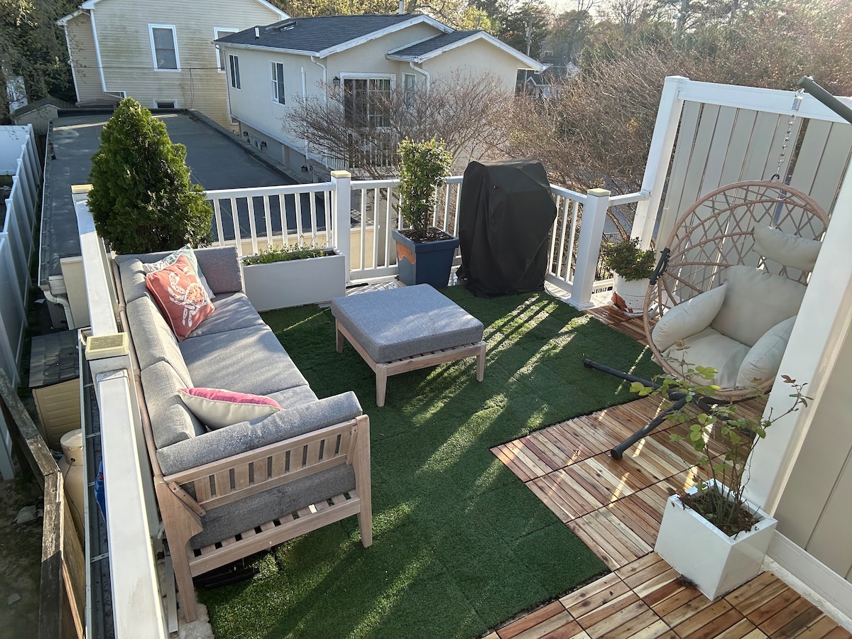 An inviting outdoor patio area is showcased, featuring a wooden seating arrangement with a gray cushion set. A small coffee table sits between the sofa and a single chair. Greenery is present in potted plants, and a grill is positioned nearby, surrounded by artificial grass.