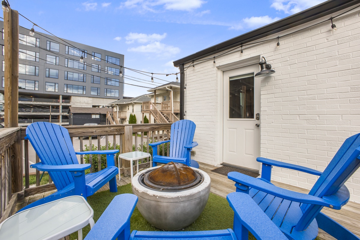 A charming outdoor space is equipped with blue Adirondack chairs surrounding a circular fire pit. A small table accompanies the seating area, while green turf adds a touch of nature. The backdrop features a modern building, creating a pleasing urban atmosphere.
