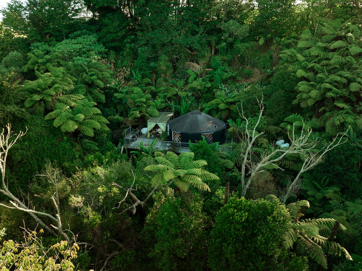 A yurt rests among lush greenery and trees, surrounded by a vibrant landscape. The expansive deck is visible, providing a natural setting that blends with the environment. The structure features a distinctive black dome shape, reminding of a cozy retreat in nature.