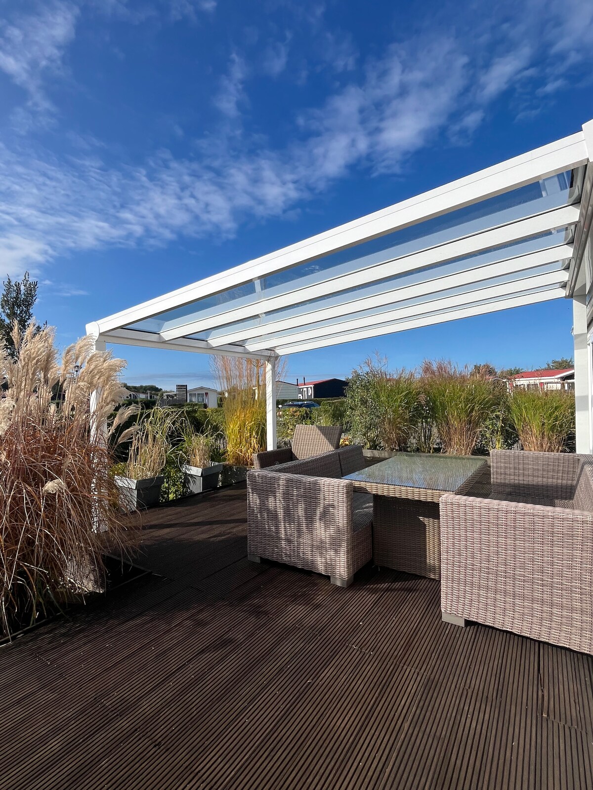 A covered terrace is visible, featuring a glass top table surrounded by a rattan seating arrangement. The area is adorned with tall grasses and shrubs, offering a serene outdoor space under a clear blue sky.