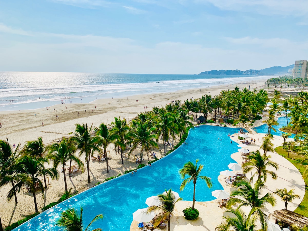 An expansive view of a turquoise pool flows alongside a sandy beach lined with palm trees. The ocean shimmers under sunlight, gently lapping at the shore where people can be seen enjoying the beach. Green hills are visible in the distance.