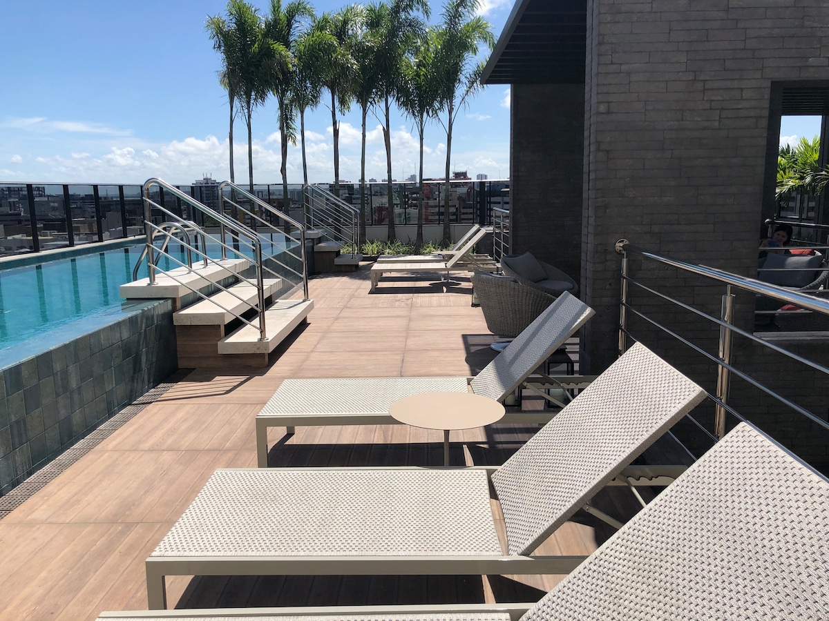 An outdoor terrace features several lounge chairs arranged around a small table, ideal for relaxation. A swimming pool is visible in the background, complemented by tall palm trees that add a touch of nature. The expansive city skyline is seen in the distance under a clear blue sky.