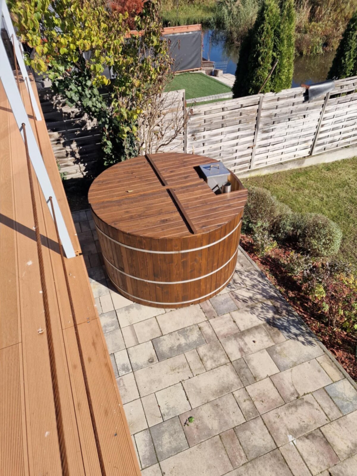 A wooden hot tub is positioned on a stone patio, surrounded by neatly trimmed grass and decorative shrubs. The area is bordered by a low wooden fence, while trees are visible in the background, contributing to a serene outdoor environment.