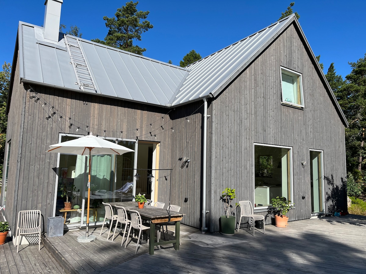 A modern wooden villa showcases a spacious outdoor terrace surrounded by nature, furnished with a large dining table and umbrella. Large glass doors allow for natural light to fill the interior, while the sloped roof and wood siding contribute to the contemporary design.