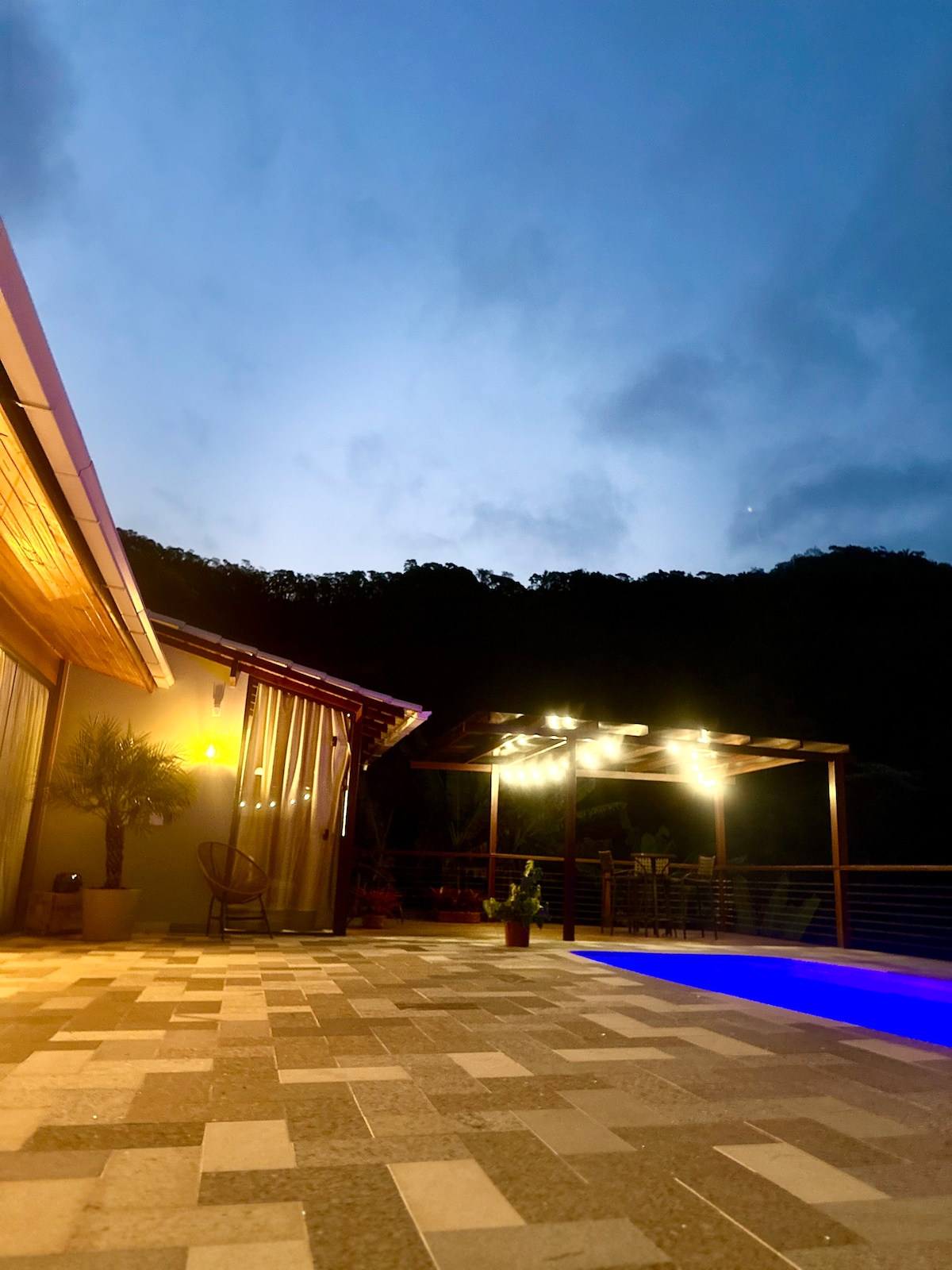 An evening view captures a serene outdoor space featuring a pool illuminated with soft blue lighting. The patio is adorned with patterned stone tiles, and a shaded area with a wooden structure houses lights, creating a welcoming ambiance against the backdrop of gentle hills.