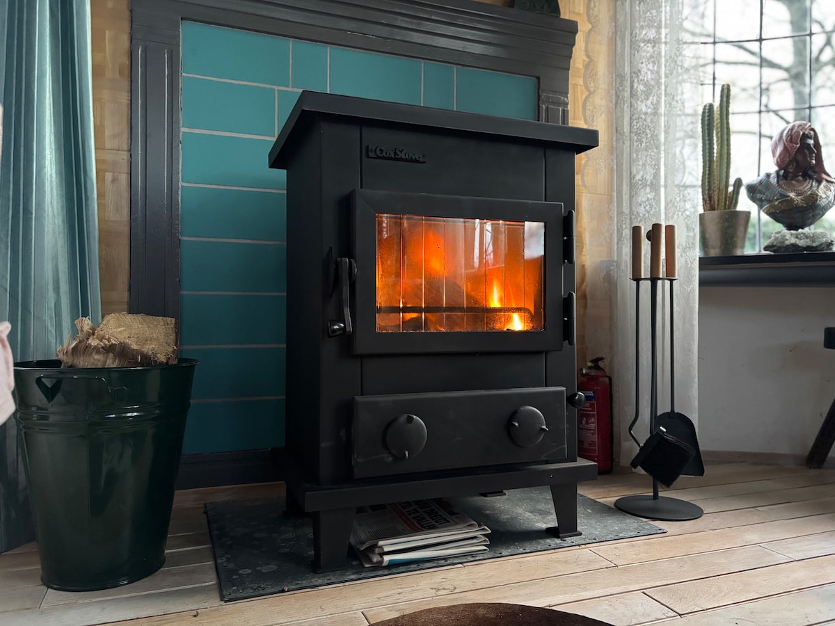 A black gas stove provides warmth, situated in front of a teal-tiled wall. The stove's glowing flames create a cozy focal point in the room. A metal firewood holder and other tools are positioned nearby, contributing to the functional ambiance of the space.