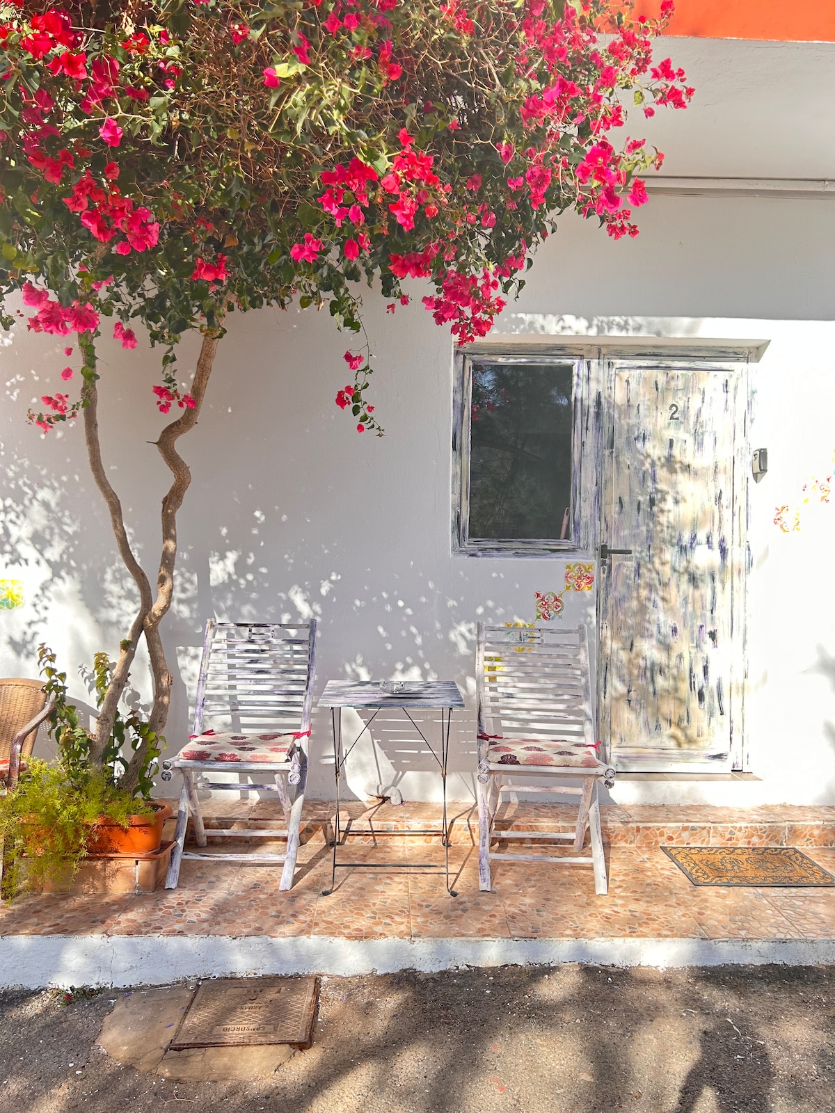 An outdoor patio area is depicted with two white wooden chairs and a small table, complemented by vibrant pink bougainvillea cascading above. The entrance to the apartment is visible, featuring a weathered wooden door and decorative wall accents.