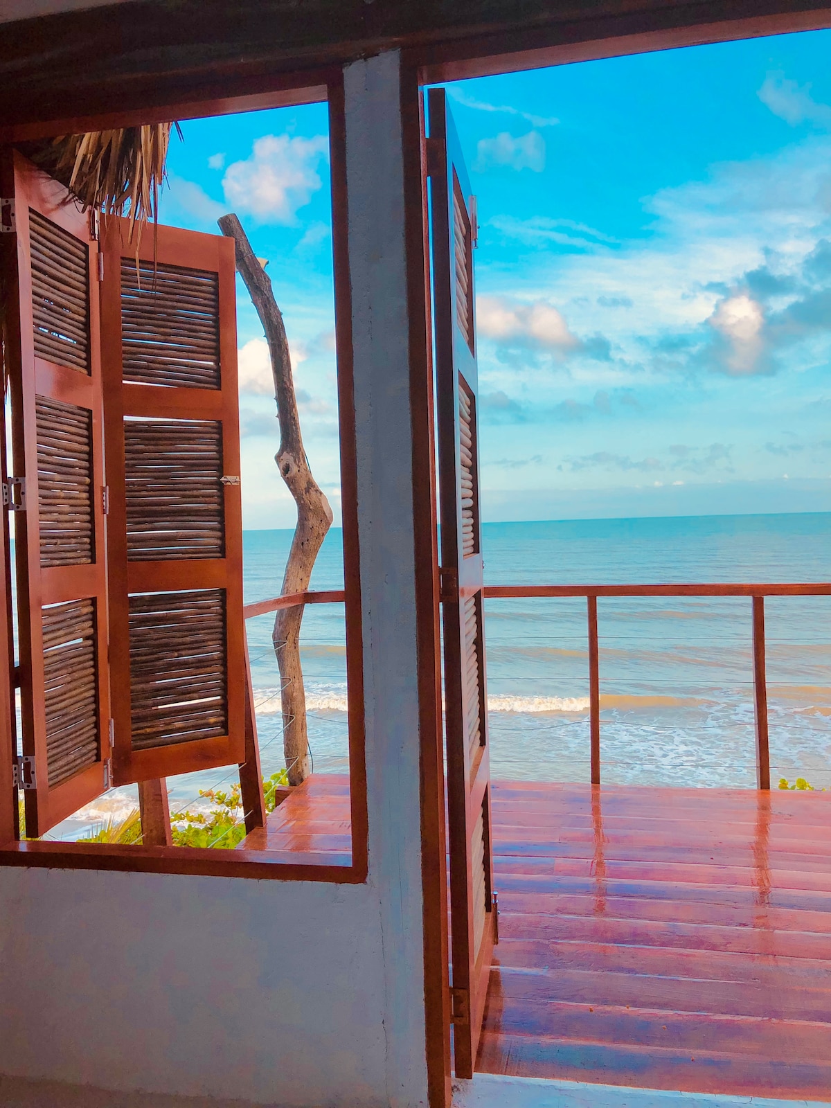 A set of wooden shuttered doors opens to reveal a scenic view of the ocean. The water reflects the sky's blue hues, while gentle waves lap at the shore. A small balcony area is visible, merging indoor comfort with an outdoor experience.
