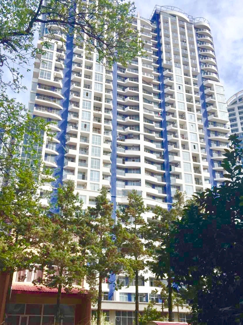 A modern residential building with a sleek design is visible, featuring multiple floors and large windows. The exterior is accented with blue and white, surrounded by green trees that lend a natural element to the urban environment.