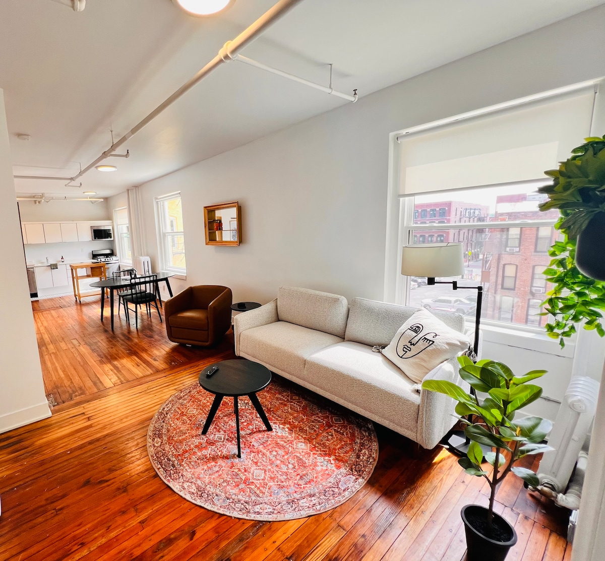 A well-lit living area features a modern sofa and armchair, complemented by a round coffee table atop a decorative rug. Large windows allow natural light to brighten the space, which includes a dining table visible in the background, highlighting the open layout.