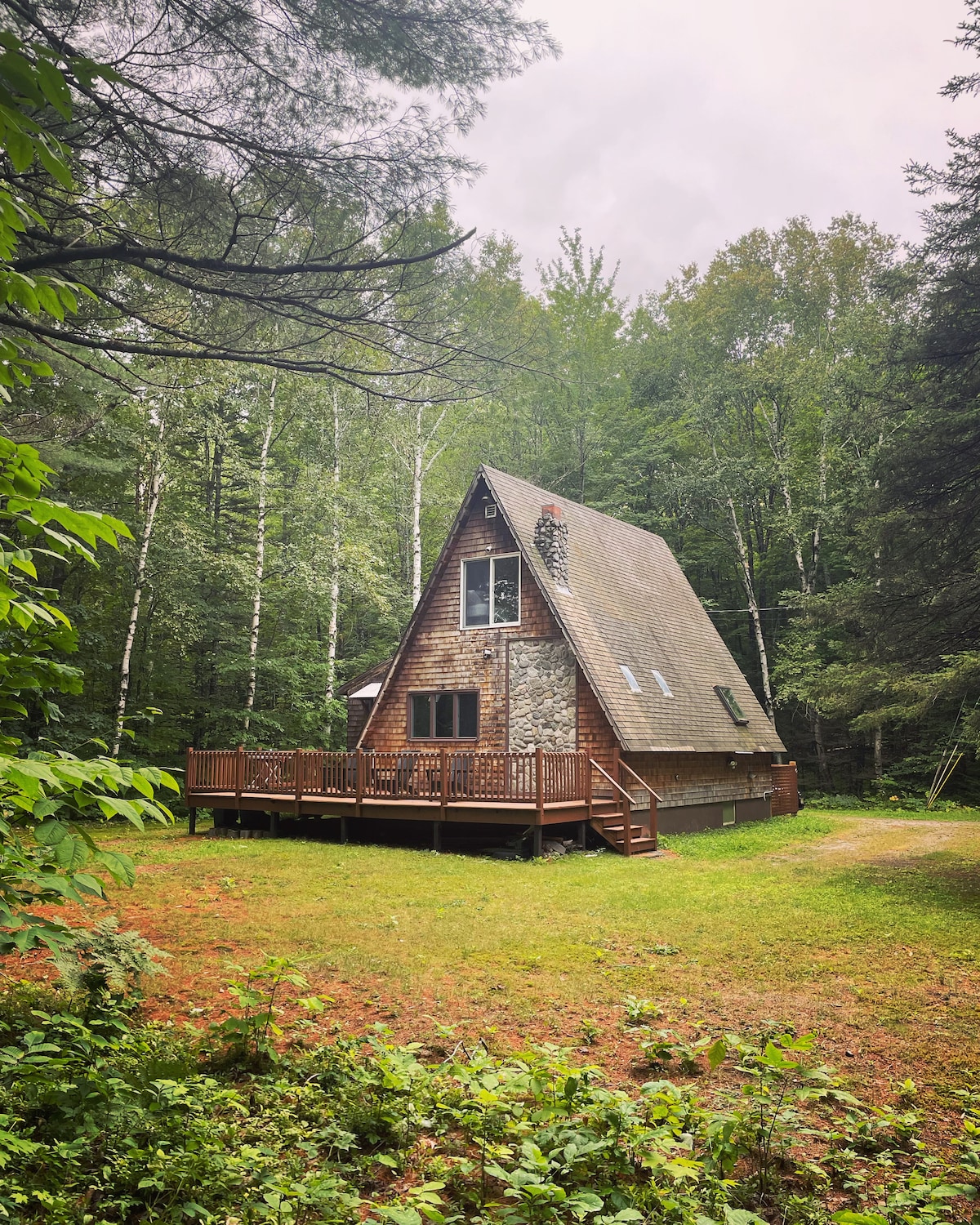 An A-Frame cabin is nestled among lush trees, showcasing a large wooden deck that extends from the front. The exterior is characterized by a mix of wood and stone, with a steeply pitched roof. Surrounding natural greenery provides a sense of privacy.