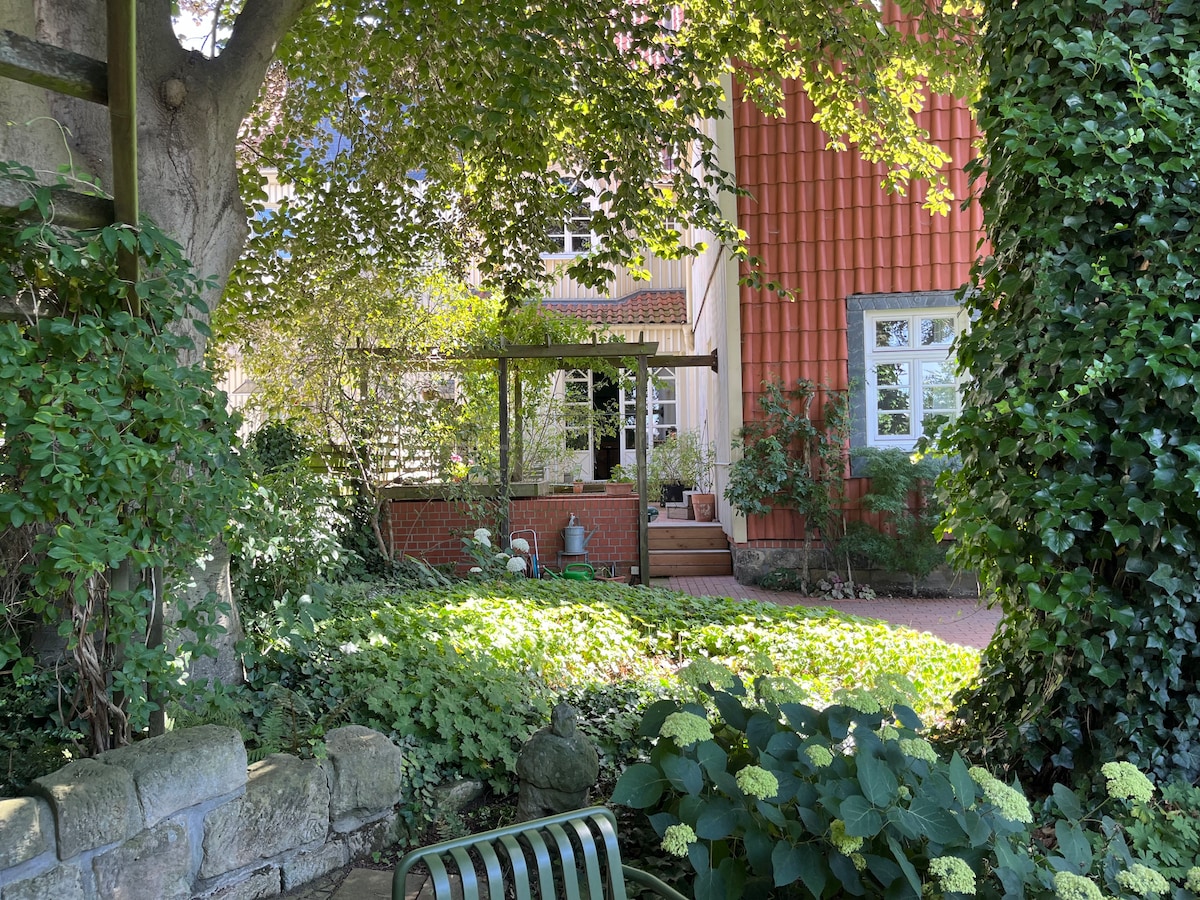 A tranquil garden scene features lush greenery with a variety of plants surrounding a small wooden deck. The red-clad exterior of the historic building provides a warm backdrop, while a shaded bench invites relaxation under the overhanging trees.