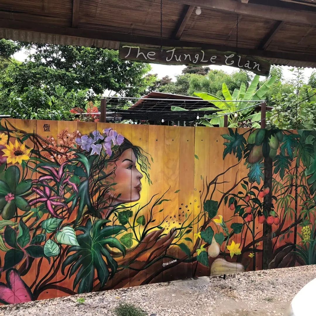 A vibrant mural featuring tropical plants and flowers is displayed on a wooden wall. The artwork incorporates various shades of green, alongside bright purples and yellows, blending seamlessly with the natural surroundings. The scene conveys a connection to nature and the environment.