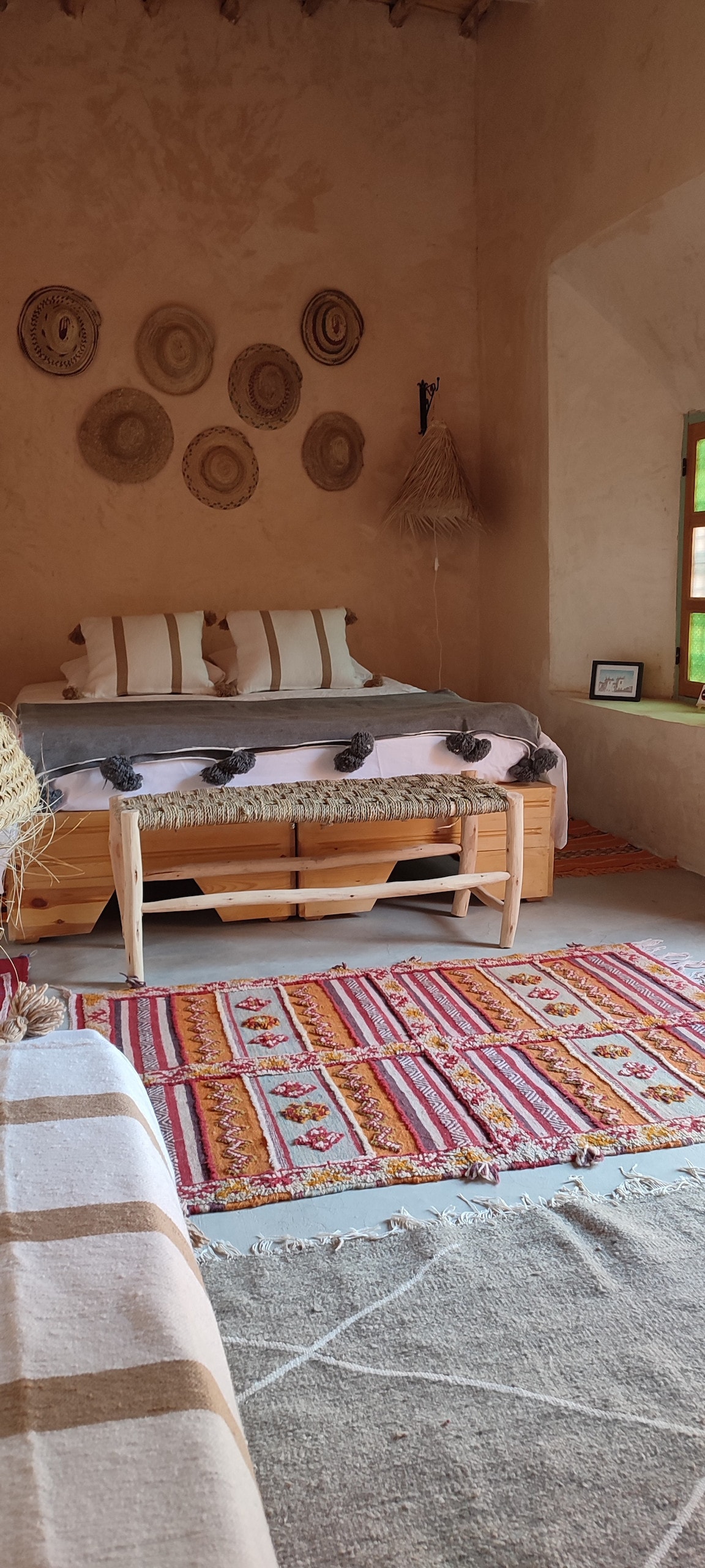 A cozy bedroom is captured featuring a spacious bed adorned with neutral-toned bedding. Decorative woven wall art adds character above the bed. A colorful area rug lies on the floor, while a window allows natural light to illuminate the room.