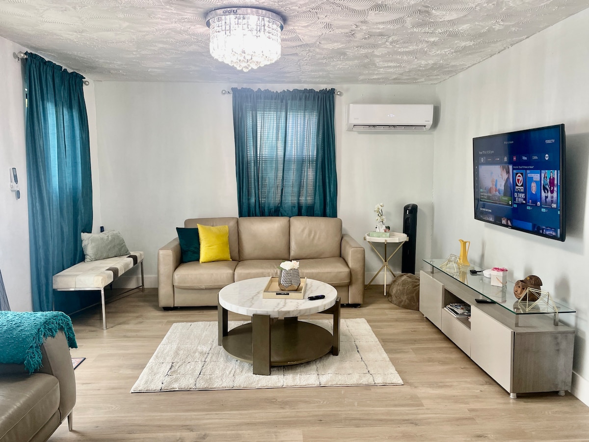 A welcoming living area is presented with a light-colored leather sofa, accented by a colorful decorative pillow. A circular coffee table sits at the center, surrounded by a cream-colored area rug. Windows are dressed in teal curtains, and a mounted television offers entertainment options.