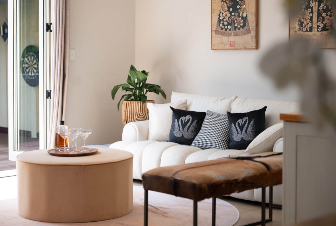 A bright living area features a cozy white sofa adorned with decorative pillows. A round coffee table sits in the center, alongside a wooden bench. Natural light floods the space through sliding glass doors, which lead outside. A potted plant adds a touch of greenery.