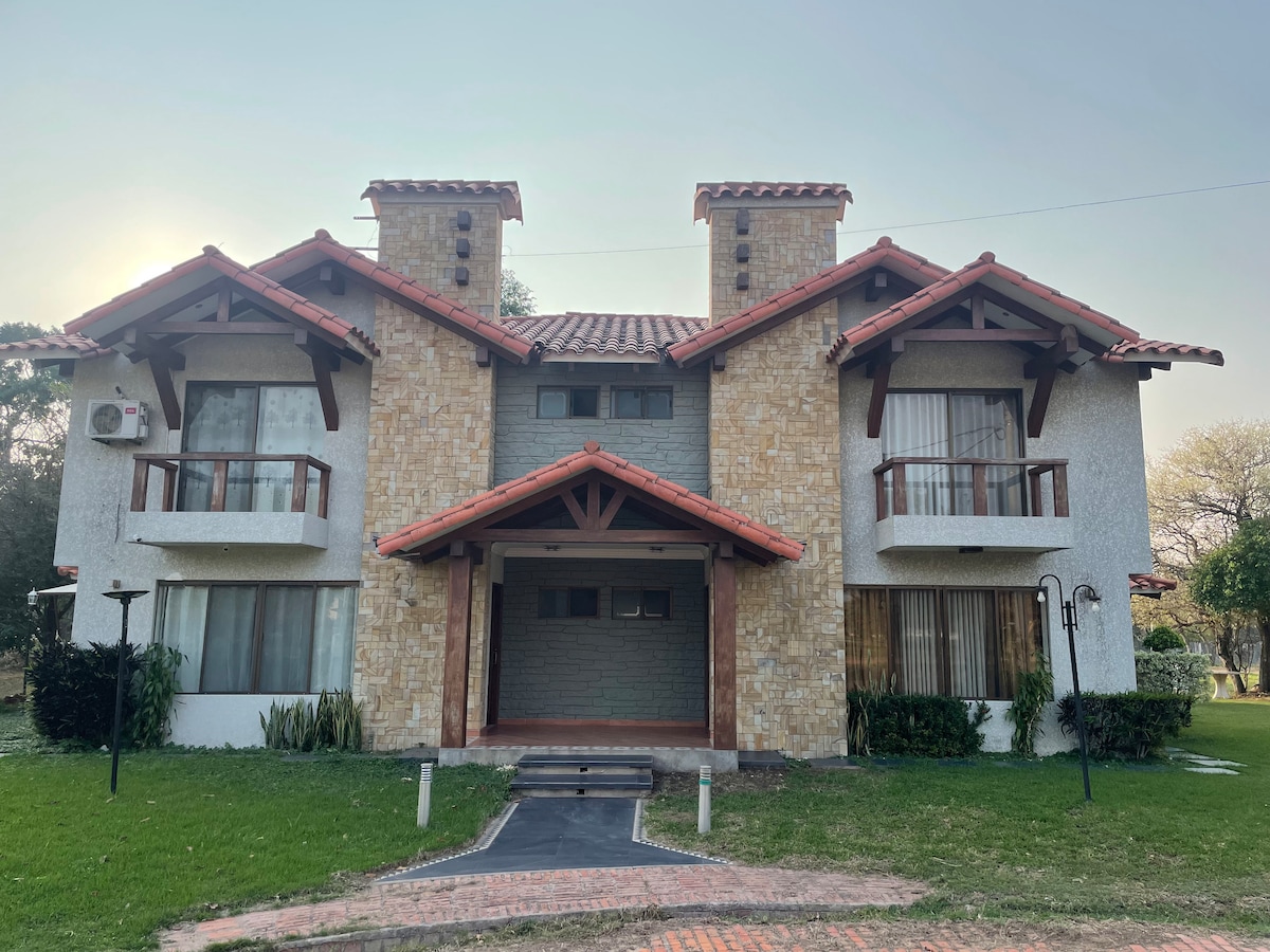 The exterior of a twin house is shown, featuring a light-colored stone facade and a red-tiled roof. Two prominent chimneys rise atop the structure, which is complemented by balconies on the upper level. Lush green grass surrounds the pathway leading to the entrance.