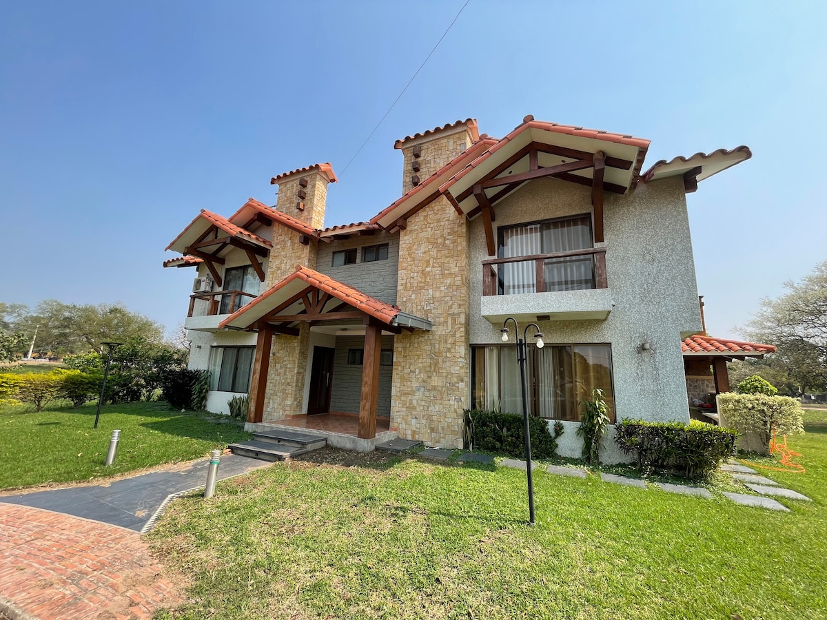 A two-story house is surrounded by lush green landscaping featuring a well-maintained lawn. The exterior showcases a combination of stone and textured finishes, with red-tiled roofs and decorative chimneys adding character. Large windows allow natural light to fill the interior spaces.