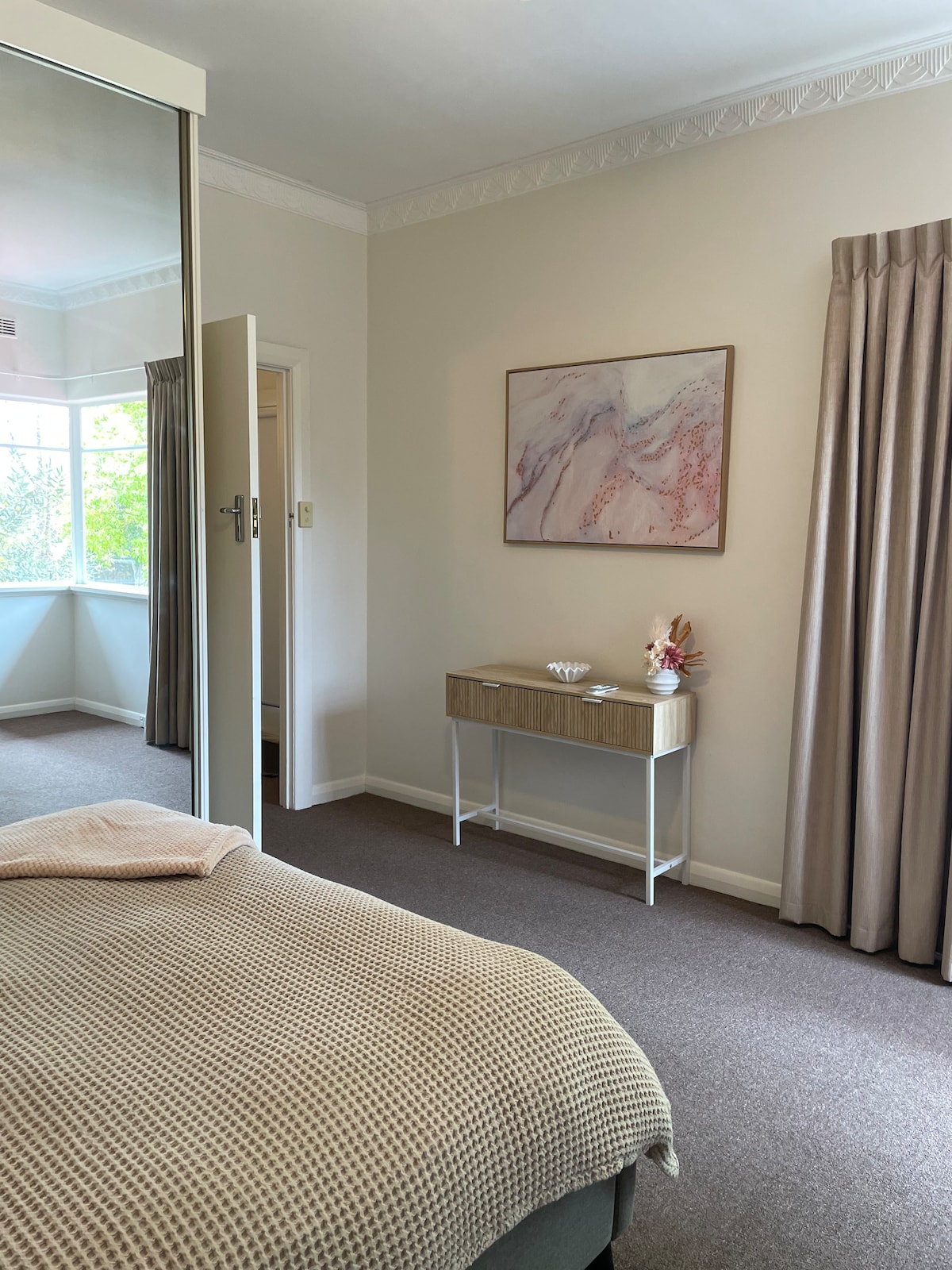 A serene bedroom setting features a cozy bed covered with a textured blanket. Soft natural light streams through a large window, illuminating the space. A minimalist console table holds decorative items, while the room's muted color palette creates a calming atmosphere.