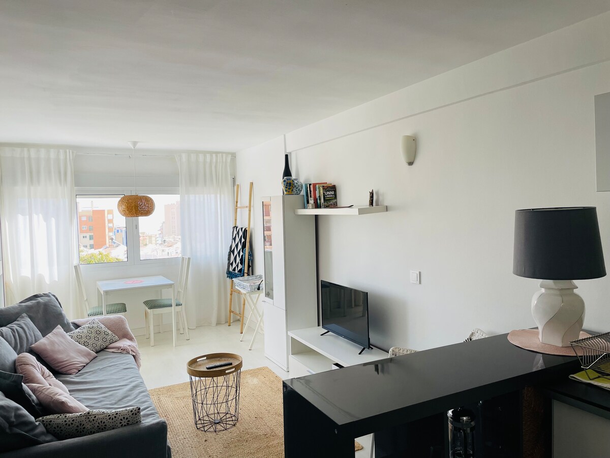 A bright living area features a spacious sofa adorned with cushions, positioned near a window dressed in sheer white curtains that allows natural light to filter in. A television is placed on a minimalistic white shelf, while a dining table is visible in the background.