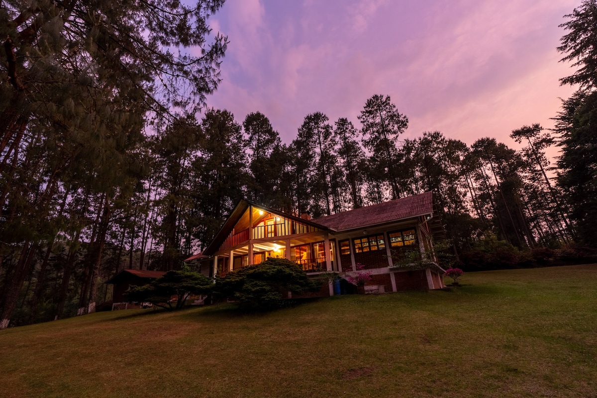 A spacious cabin is set against a backdrop of tall trees, with warm light spilling from large windows. The evening sky is painted in shades of pink and purple, creating a serene atmosphere amidst the surrounding nature.
