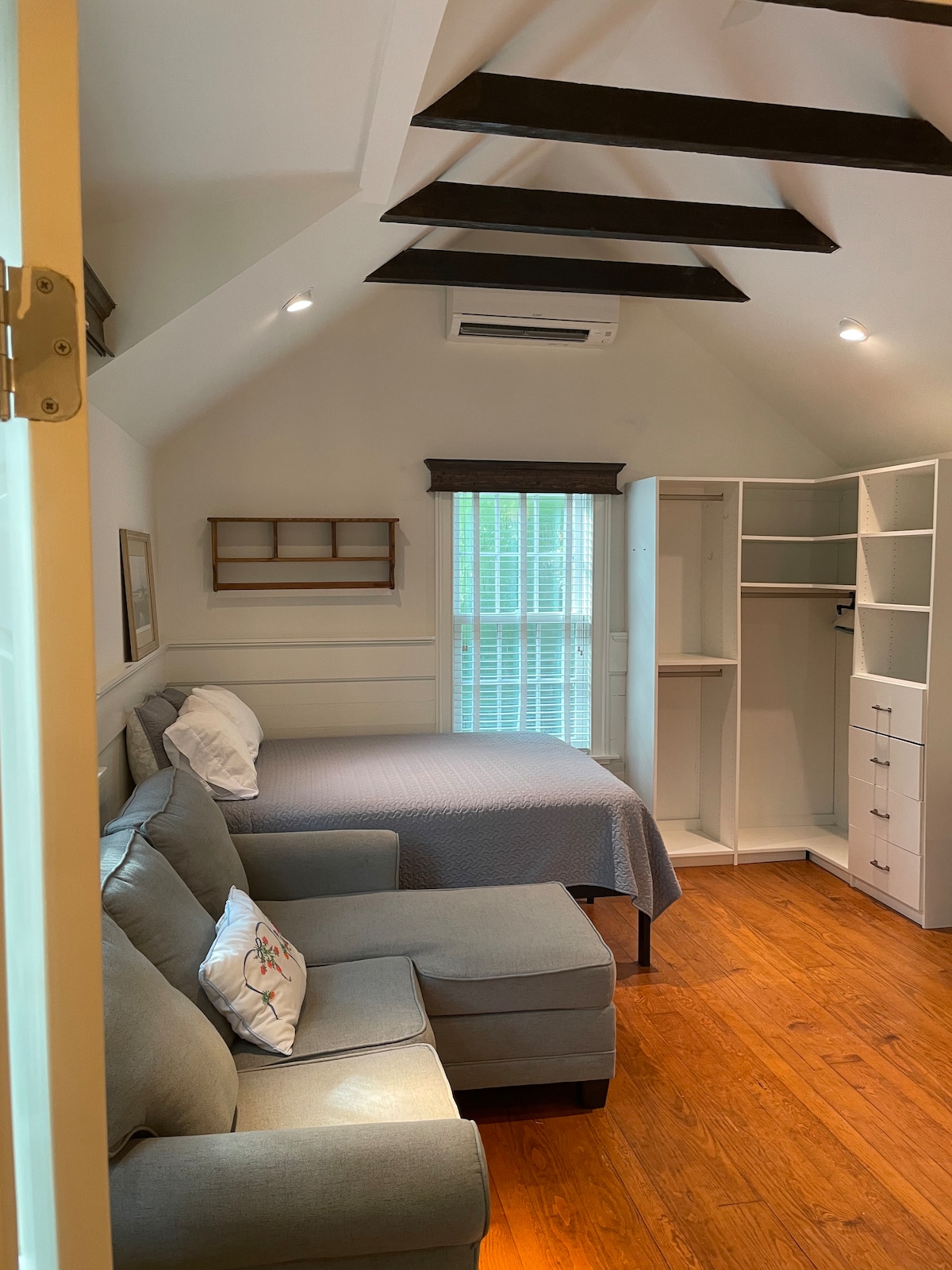A cozy studio space features a comfortable gray sofa with a decorative pillow and a queen bed draped in a soft blanket. Natural light filters through a window, illuminating the hardwood floor and exposed ceiling beams. A built-in closet system provides organized storage options.