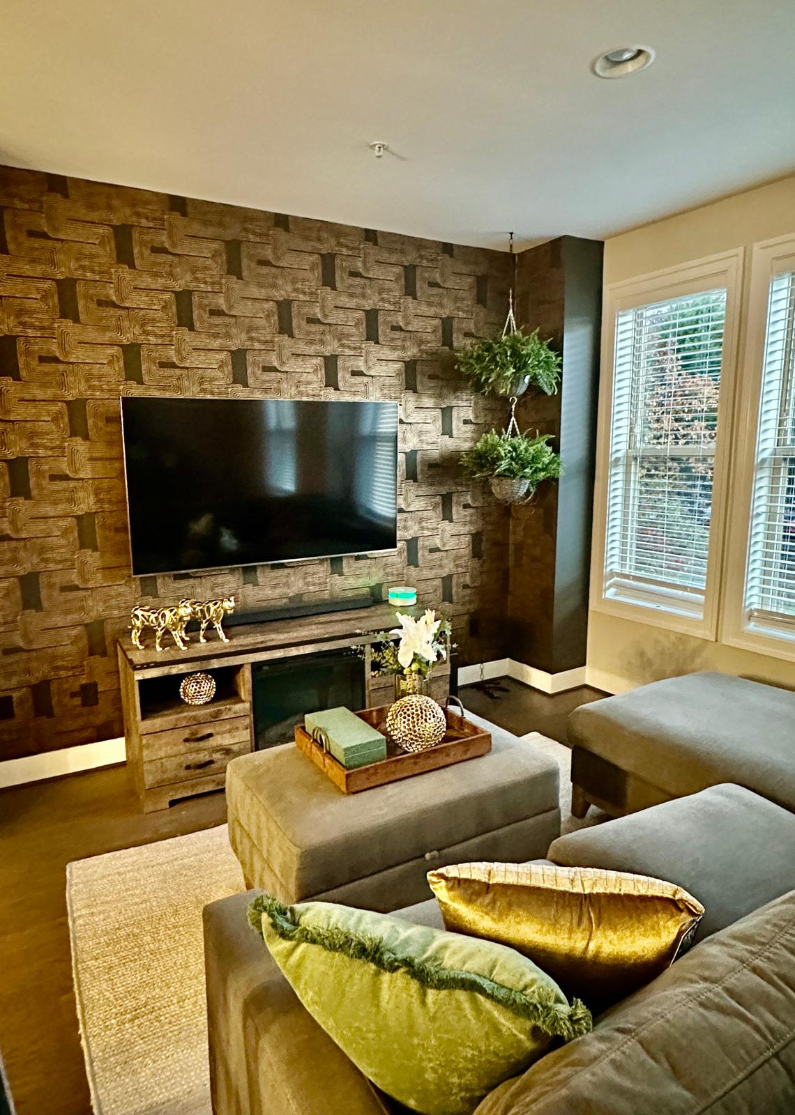 A living area is displayed with a mounted flat-screen TV on a textured wall, complemented by decorative plants. A plush sofa with colorful cushions faces the TV, and sunlight brightens the space through the large, multi-pane windows.