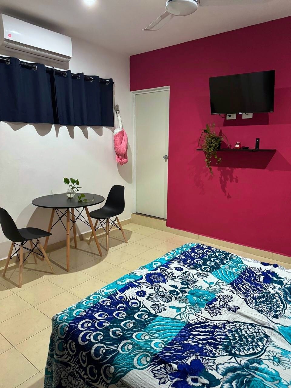 A well-lit room features a cozy sleeping area with a patterned bedspread. A small table with two chairs is positioned near a window covered by navy curtains. A wall-mounted TV and small shelf add functionality, while a door leads to another space.