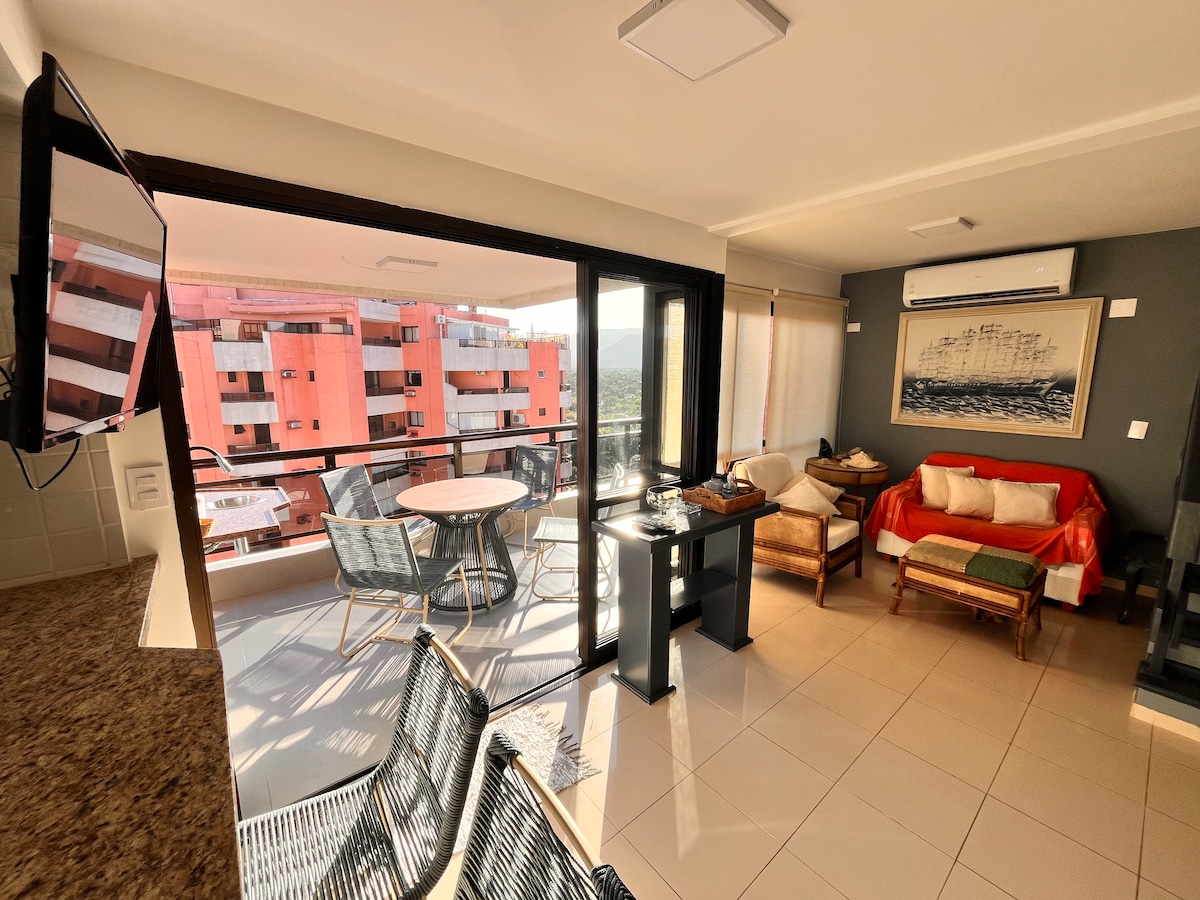 A light-filled living area features large glass doors opening to a balcony. Comfortable seating includes a red couch and wicker chairs, arranged around a small table. A media console with a television is positioned against the wall, creating an inviting space for relaxation.