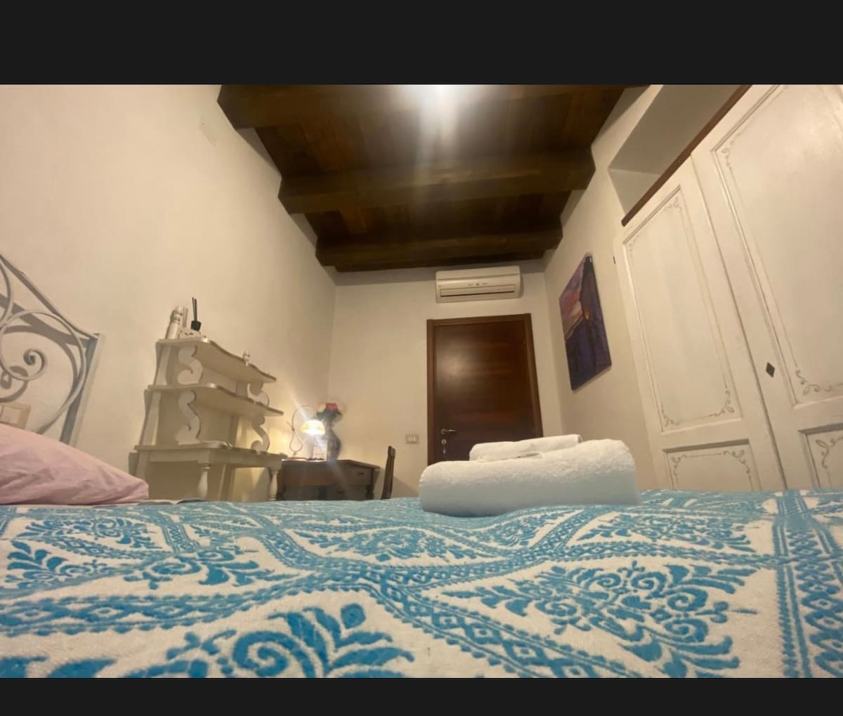 A cozy room features wooden beams on the ceiling, a spacious bed adorned with a blue and white patterned blanket, and a small desk. A soft light illuminates the space from a nearby lamp, while white cabinets provide storage options.