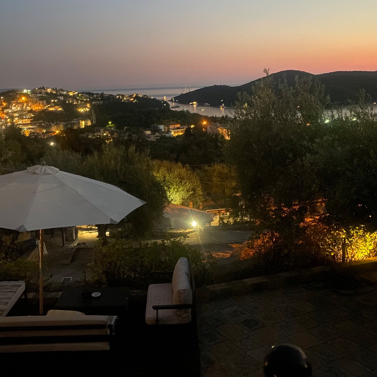 A scenic sunset view over Sivota Bay is captured, highlighting the glowing lights of the nearby village. Olive trees and an umbrella shade part of the outdoor seating area, enhancing the serene atmosphere. The coastline and distant hills create a calming backdrop.