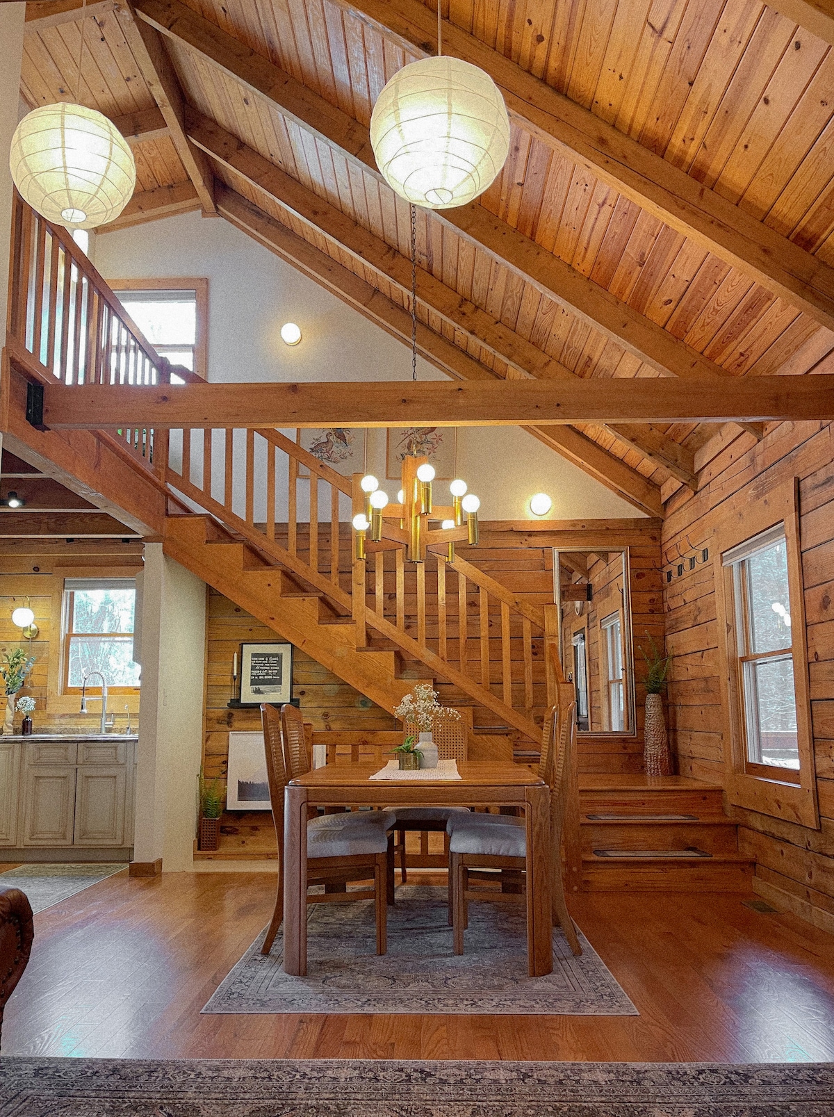 An open-concept space features a dining table surrounded by four chairs, positioned beneath a pendant light. Vaulted ceilings reveal wooden beams, while large windows allow natural light to fill the area. A staircase leads to an upper level, enhancing the sense of spaciousness.
