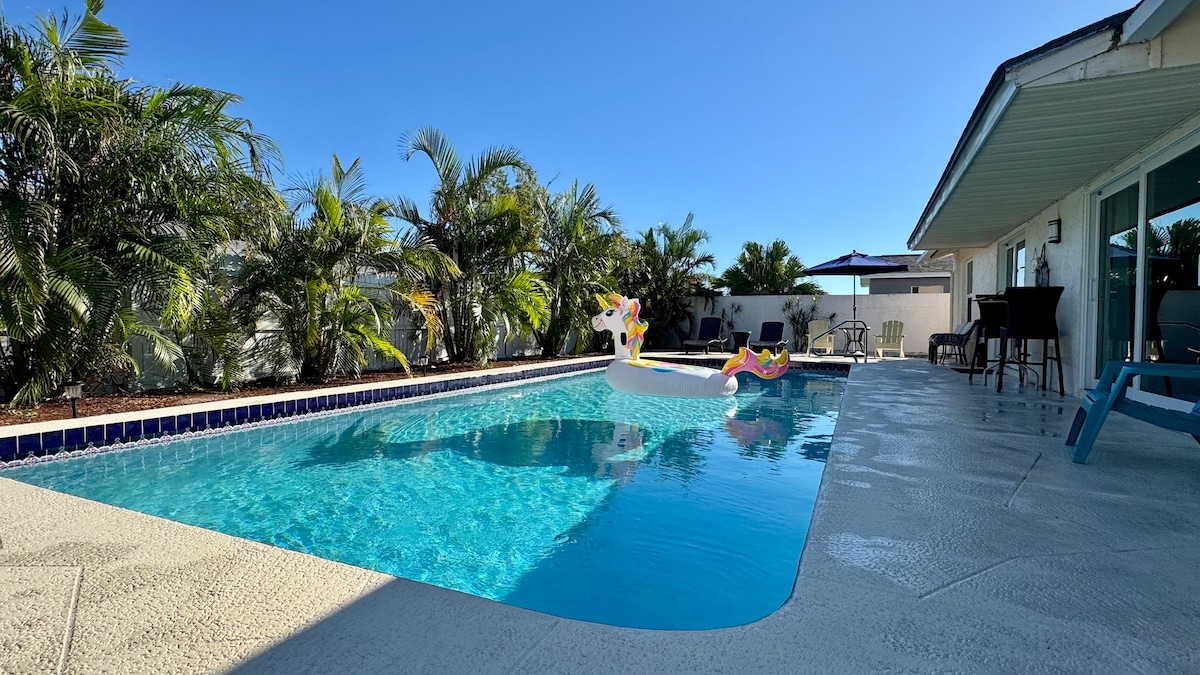 A clear, inviting saltwater pool is surrounded by lush tropical palms. A large deck area offers seating with an umbrella, while a colorful inflatable floats on the water's surface, adding a playful touch to the serene outdoor space.