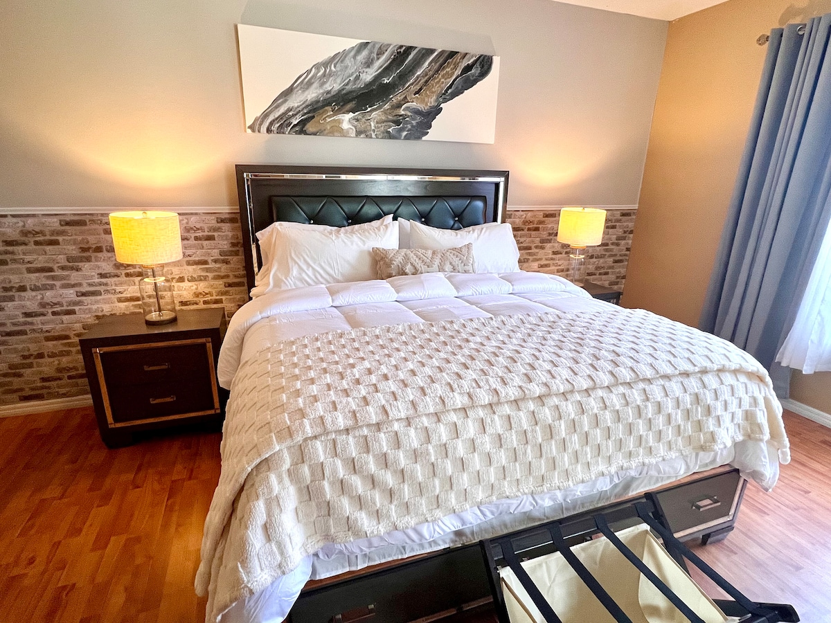 A cozy sleeping area features a spacious bed with a textured cream coverlet, complemented by two bedside lamps emitting soft light. A modern artwork piece hangs above the headboard, and light streams in through a nearby window draped with soft curtains.