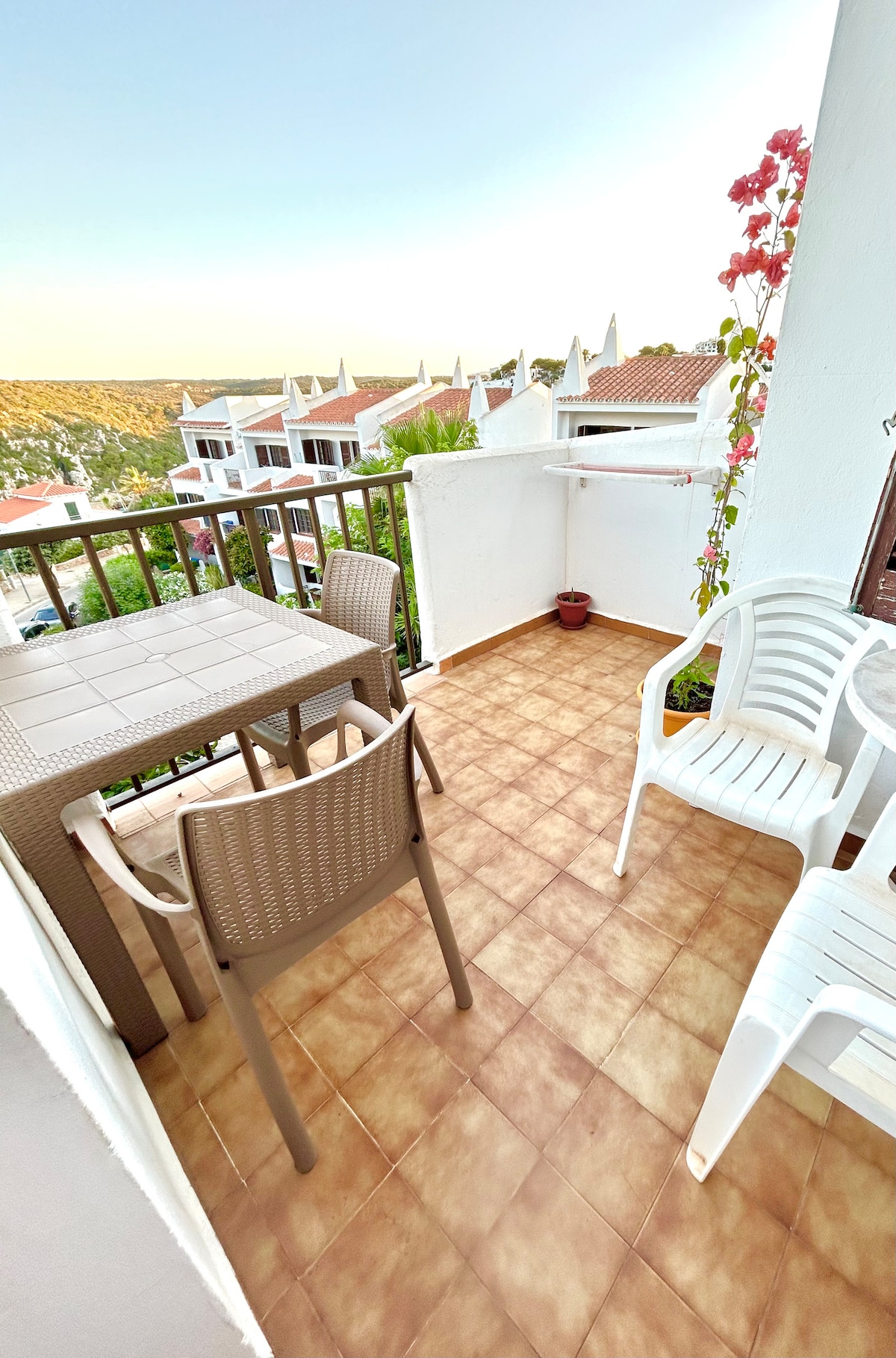 A balcony is shown with a small table and two chairs, surrounded by a railing. Potted plants add a touch of greenery. The tiled floor reflects natural light, and the view of the surrounding area is visible in the background.