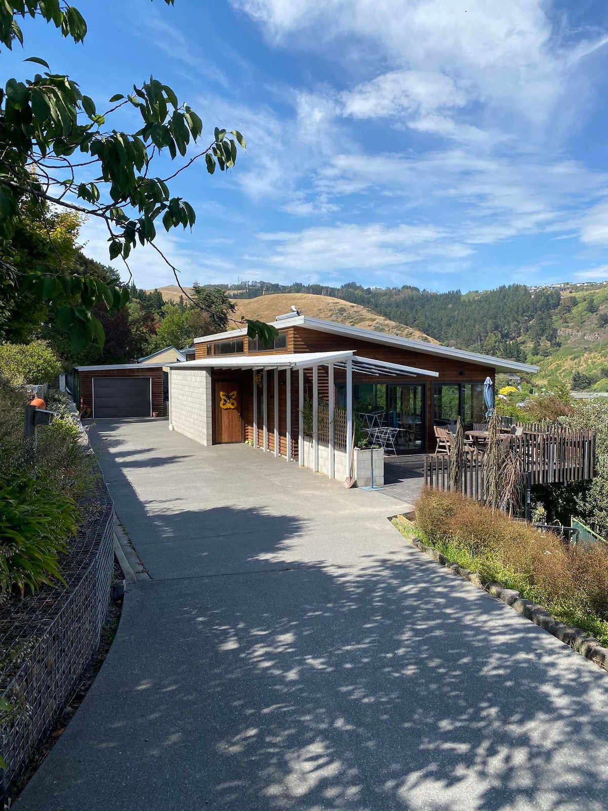 The exterior of the guest house is shown, with modern architecture featuring large windows and a wooden overhang. A pathway leads to the entrance, flanked by greenery. The surrounding hills are visible in the background under a partly cloudy sky.