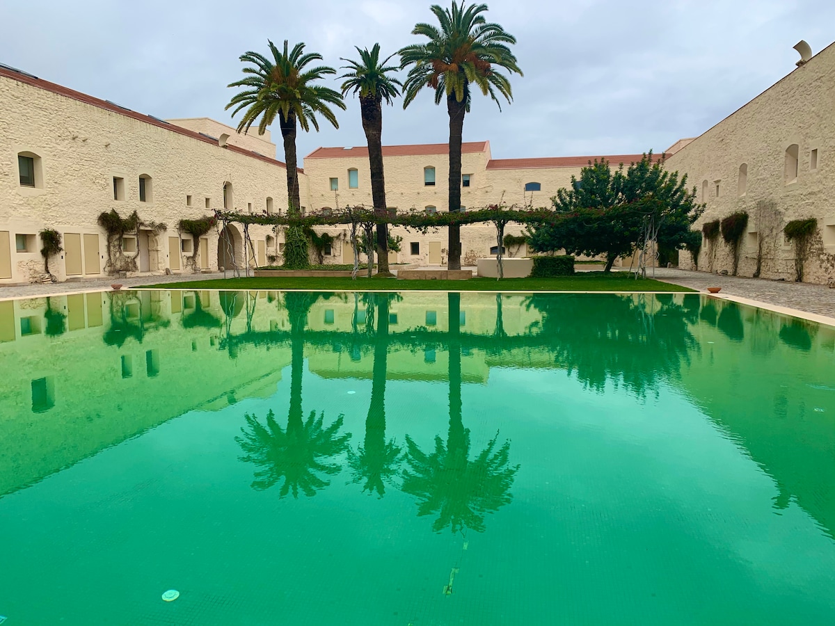 A large swimming pool is surrounded by palm trees and low stone walls, reflecting a serene atmosphere. The water appears emerald green, and the stillness of the scene adds to the tranquil environment. Soft lighting enhances the natural beauty of the outdoor area.