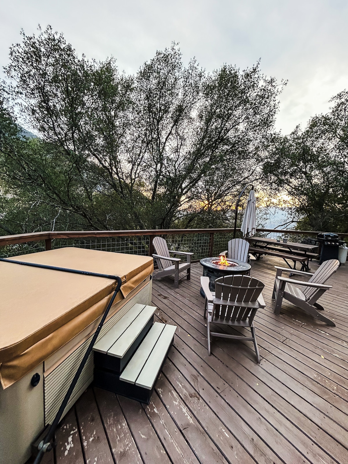 An outdoor deck features a hot tub with steps leading up to it. Surrounding seating includes several chairs and a circular table, enhancing the space for relaxation. Lush trees frame the background, complementing the serene natural setting.