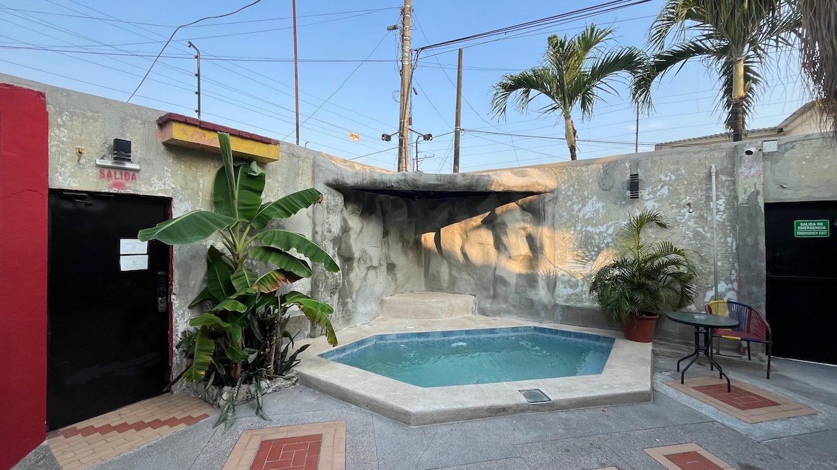 A small soaking pool is positioned within a courtyard, surrounded by tropical plants and palm trees. The area is paved with decorative tiles, and a shaded seating space is visible nearby, providing a relaxing atmosphere for guests.