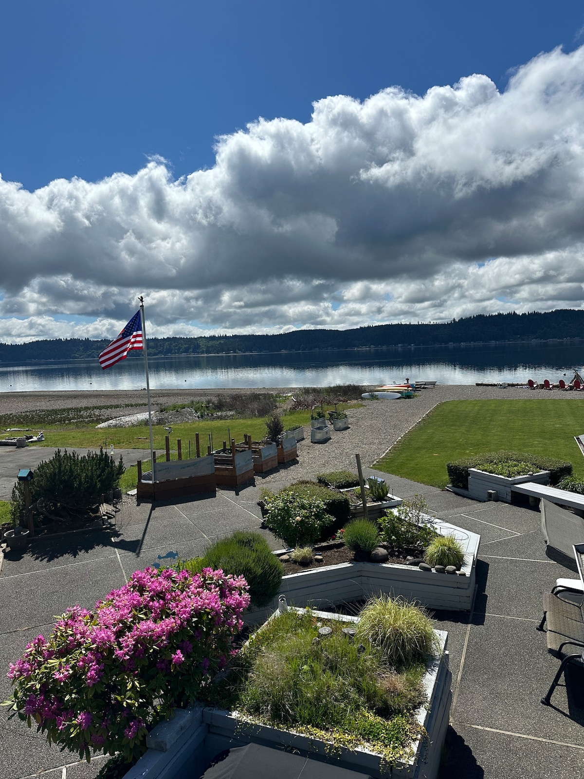 A view of the waterfront showcases a peaceful scene with a calm water surface reflecting clouds. In the foreground, colorful flowers in garden beds complement green grass and a variety of outdoor seating areas. An American flag flutters gently by the water's edge.