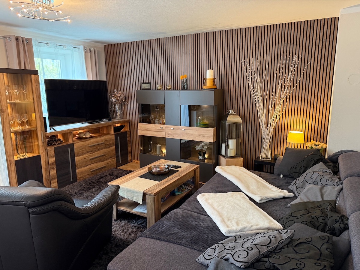 A spacious living area features a dark sectional sofa adorned with soft throw pillows. A wooden coffee table sits at the center, accompanied by an armchair. A large television is mounted on the wall opposite, while decorative shelves house various items, creating a warm ambiance.
