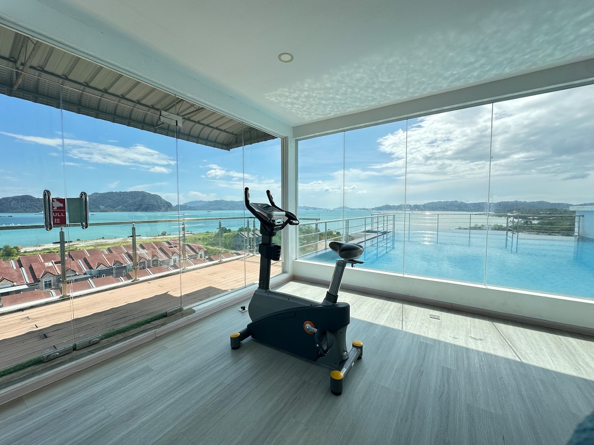 A fitness area is equipped with a stationary bike, featuring floor-to-ceiling glass walls that provide a panoramic view of the nearby coastline and landscape. Natural light brightens the space, creating an inviting environment for exercise.