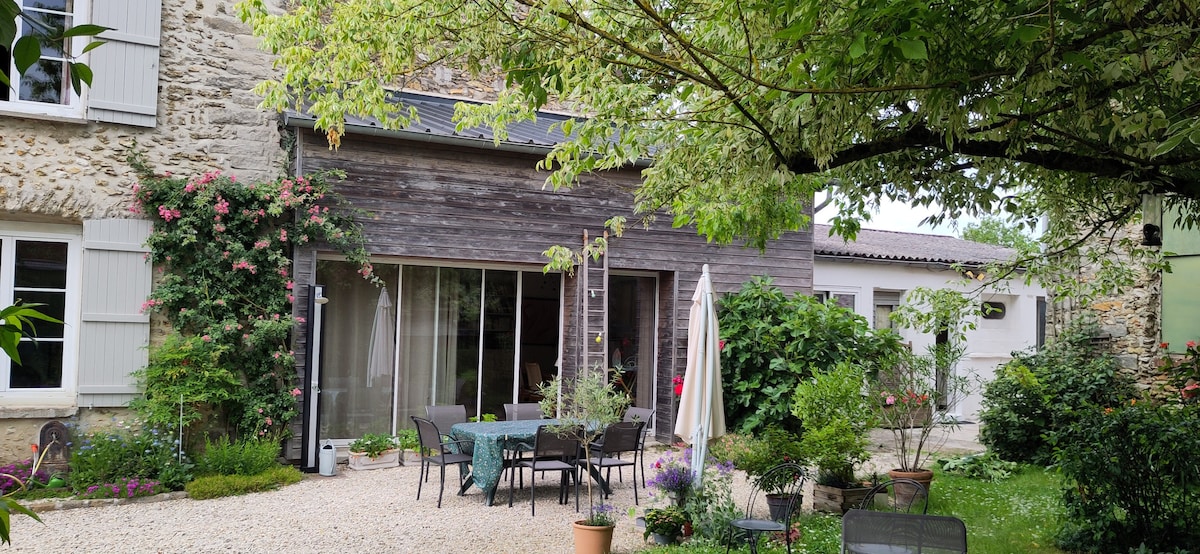 A spacious outdoor area is framed by lush greenery, featuring several tables and chairs arranged on gravel. A large umbrella provides shade, while potted plants add color to the setting. The wooden structure in the background highlights the natural blend of indoor and outdoor living.