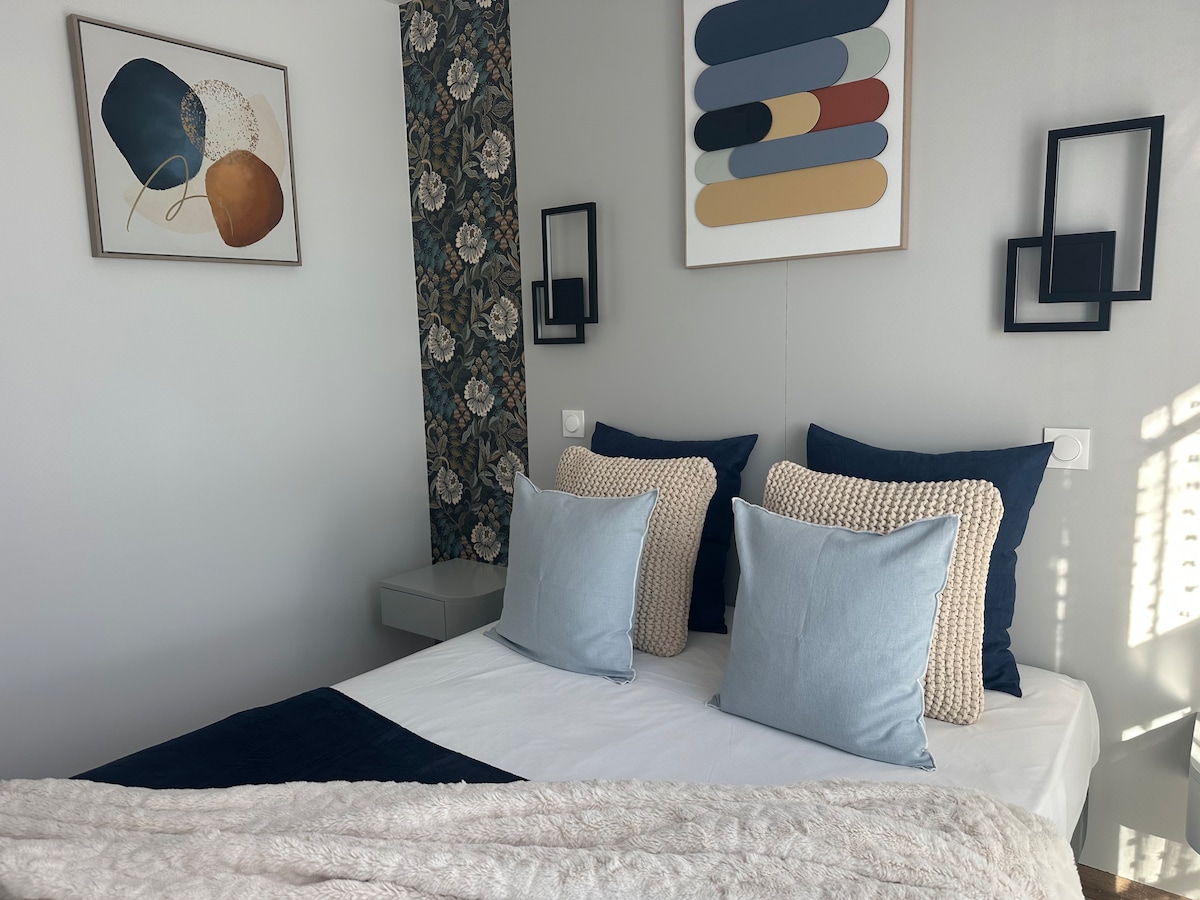 A cozy sleeping area features a double bed adorned with soft blue and cream pillows. Elegant wall art is displayed above the bed, complemented by decorative wallpaper. Natural light filters through the window, enhancing the soothing color palette.