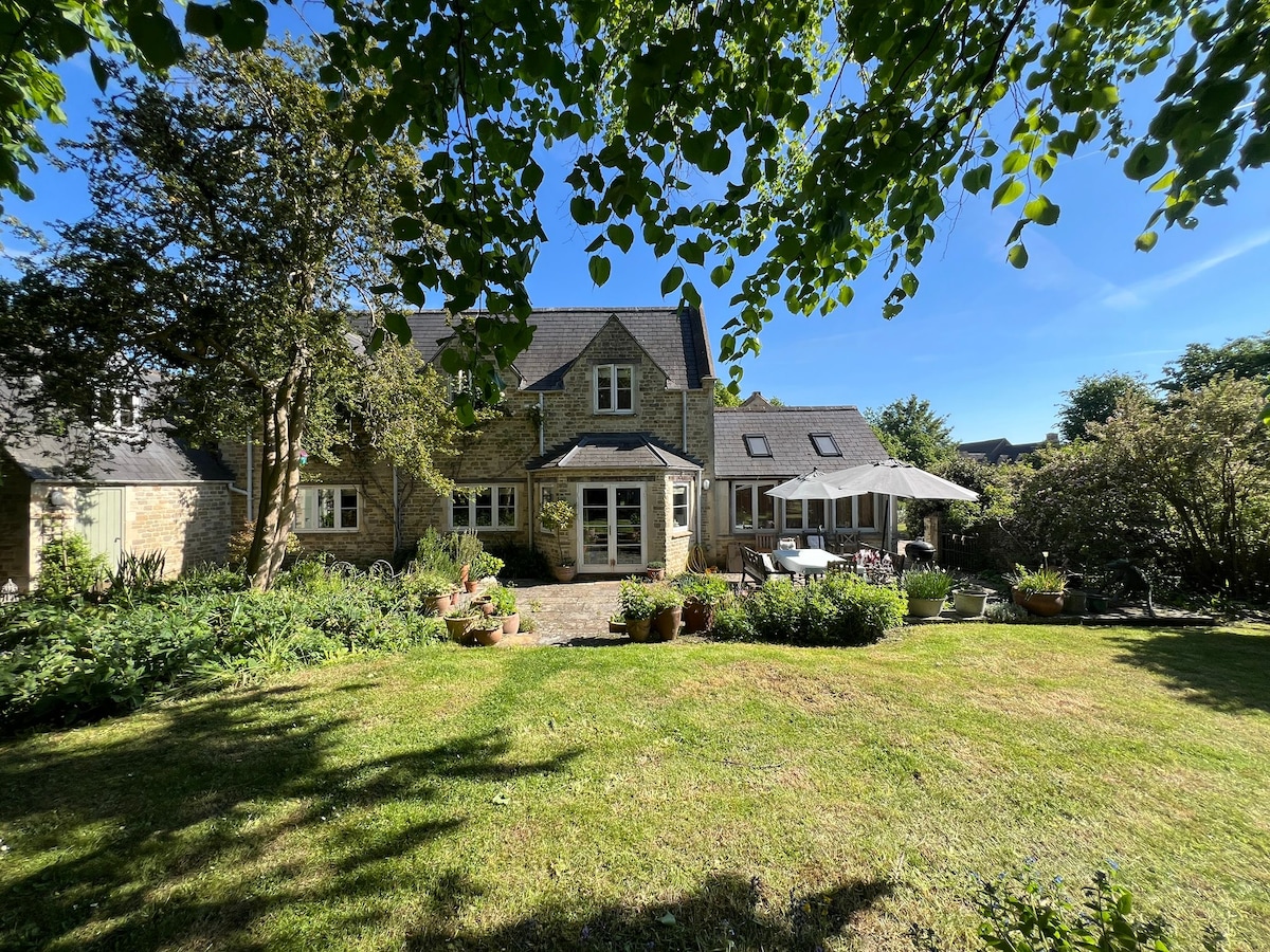 A spacious garden is visible, featuring a well-maintained lawn surrounded by various plants and pots. The cottage's exterior showcases charming stonework with windows that allow natural light. A patio area is covered by an umbrella, offering a shaded spot for outdoor gatherings.